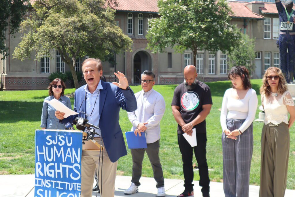 Fair-skinned man wearing a blue blazer and blue button down shirt speaking at a podium reading "SJSU Human Rights Institute Silicon" in front of six people near lawn