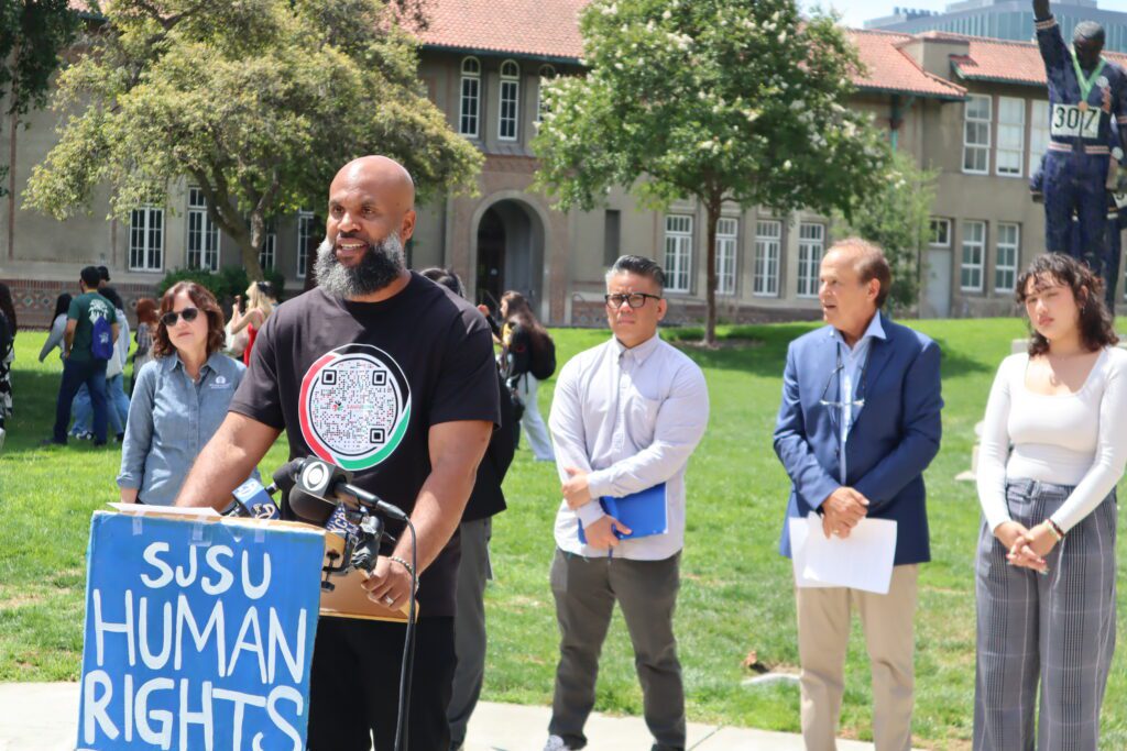 Bald, dark-skinned man with a beard wearing a black t-shirt with a QR-code speaking at podium with sign reading "SJSU Human Rights" in front of five other people