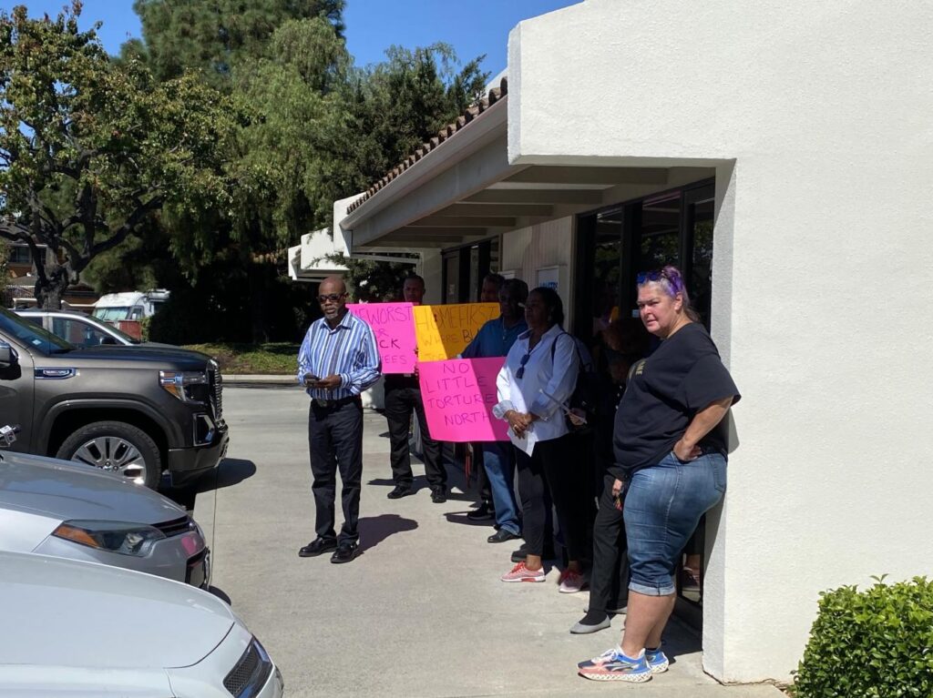 Rev. Jethroe Moore II standing on the sidewalk in front of HomeFirst's offices, alongside former Sunnyvale Family Shelter employees who are shaded by the building's roof.