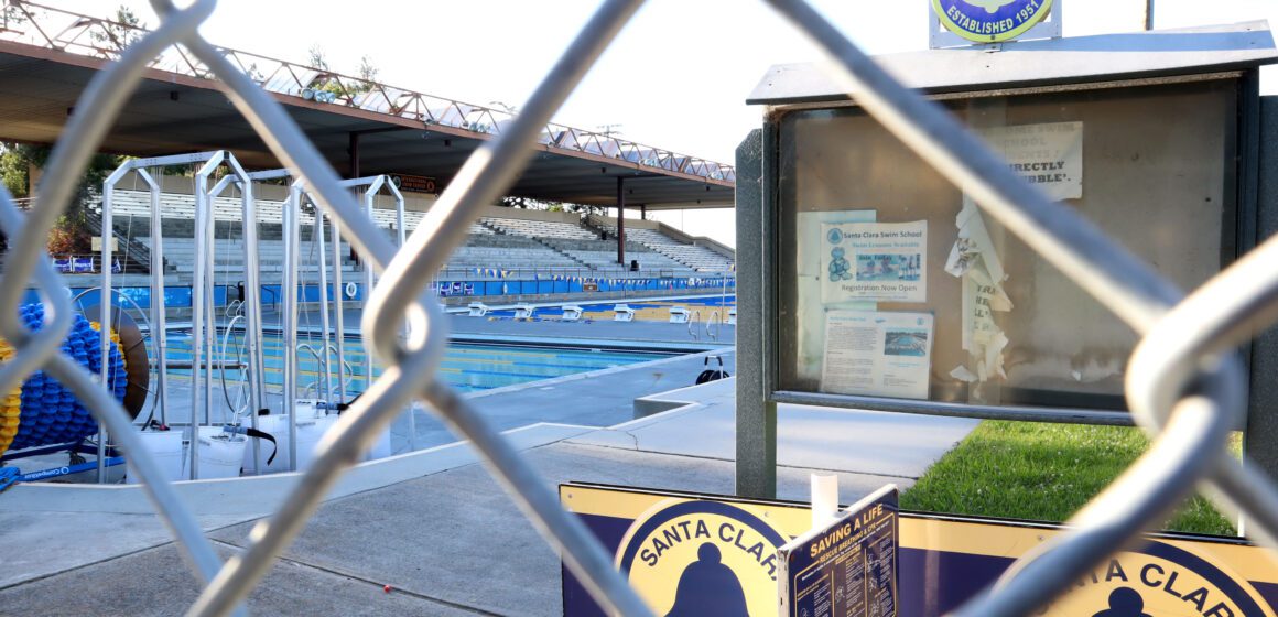 Chain link fence in front of the Santa Clara Swim Club's empty notice board and two large pools with an empty audience stands.