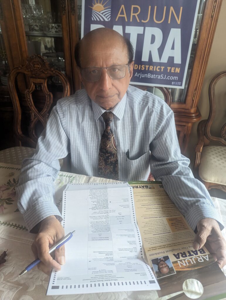 An elected official sits at a table and completes his ballot while a campaign sign appears behind him,