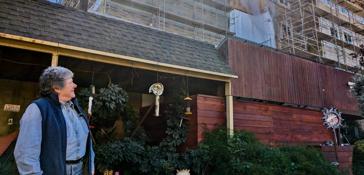 A woman in her front yard looks at a six-story apartment tower being constructed behind her house in San Jose.