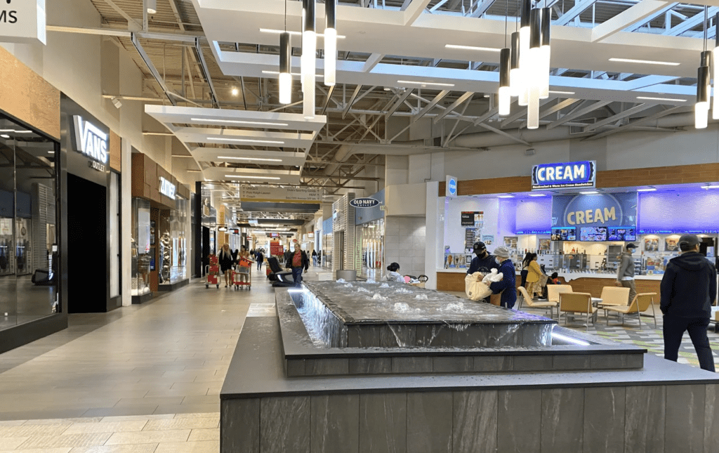 Inside Milpitas Great Mall, looking down hallway at shoppers sitting and standing by indoor water feature