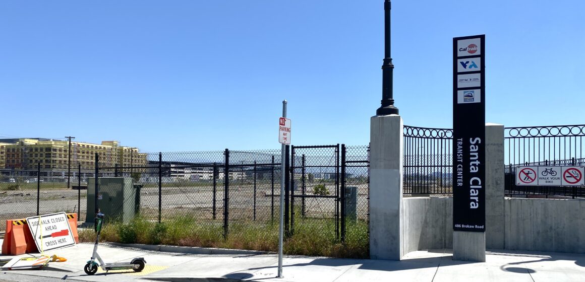 The Santa Clara Transit Center sign by the sidewalk, with a fenced-in open plot of land