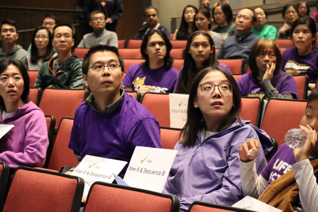 Fremont Union High School District parents wearing purple sit in an auditorium holding signs that read map six and sequence b with a check mark.