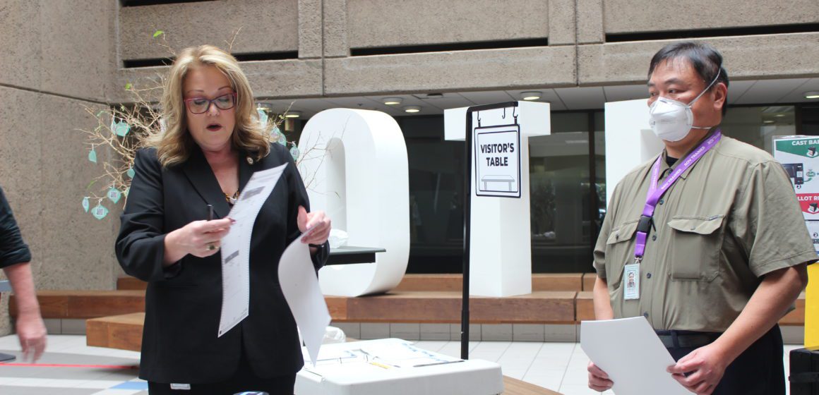 A woman stands on the left with a voter ballot and a man stands on the right holding a clipboard