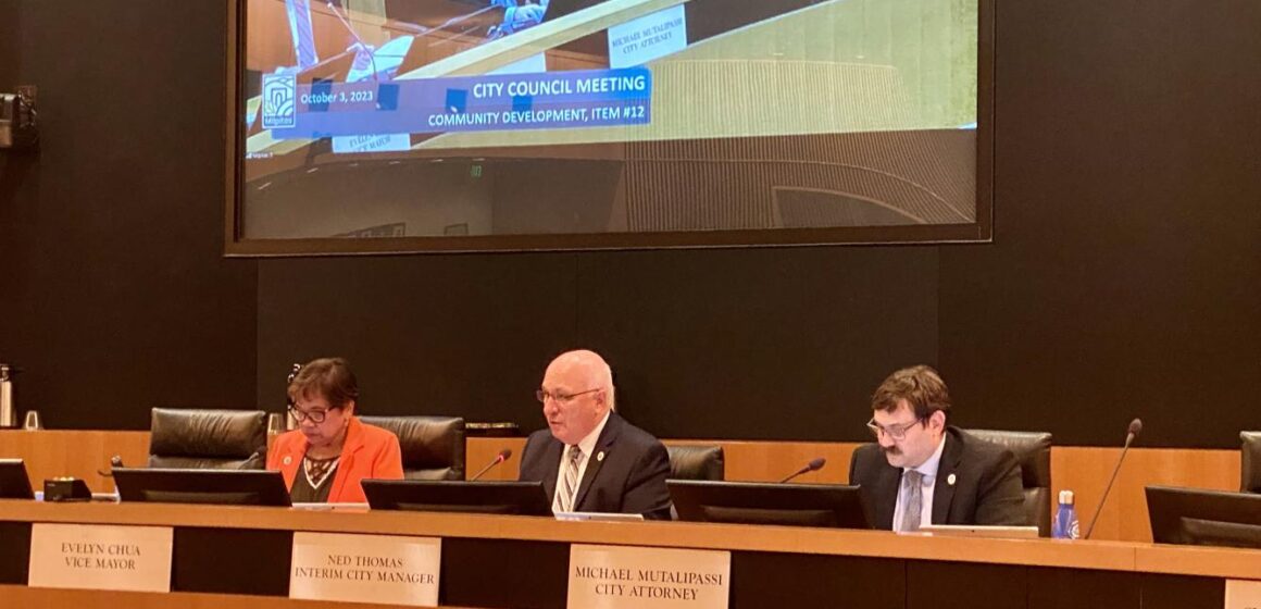 Milpitas interim city manager Ned Thomas speaking and sitting behind the city council dias. City attorney Michael Mutalipassi and Vice Mayor Evelyn Chua sit beside him.