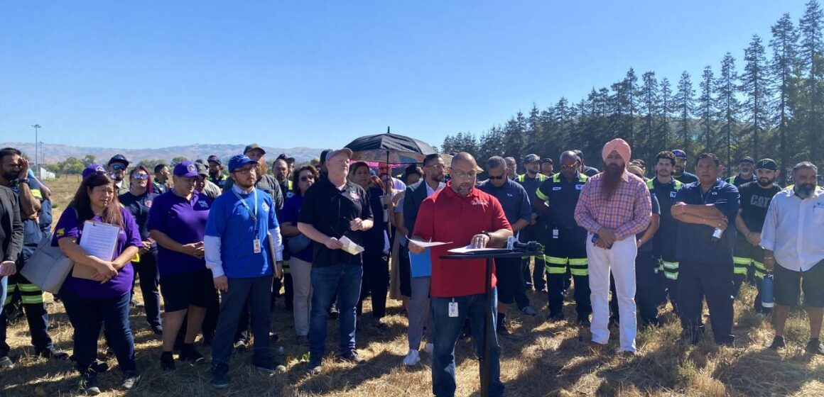 AFSCME Local 1101 President Steve Jovel speaks in front of a crowd of about 50 at the VTA Cerone Yard, standing at a podium over a field of dried grass.