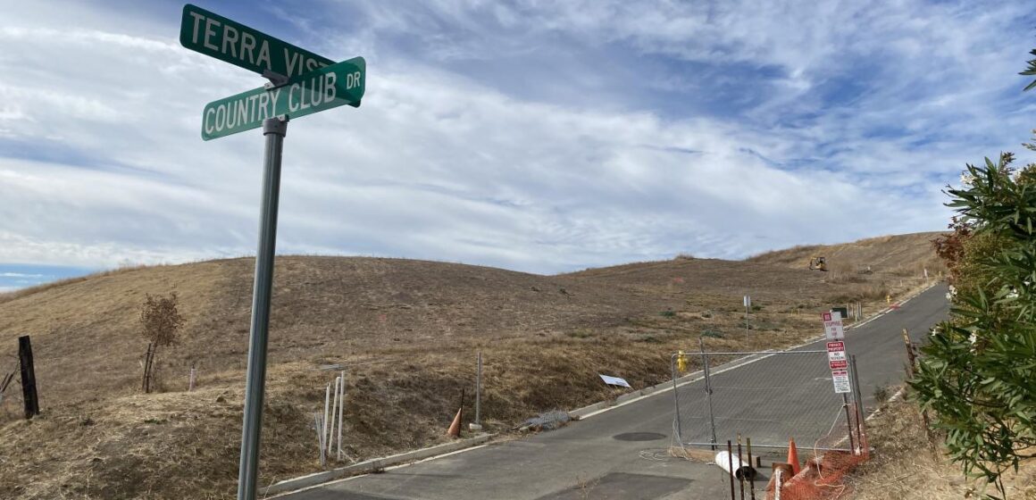 Intersection of Terra Vista Court and Country Club Drive, where multiple empty lots of grassy hills sit along the street, which is partially fenced off with construction equipment in the distance