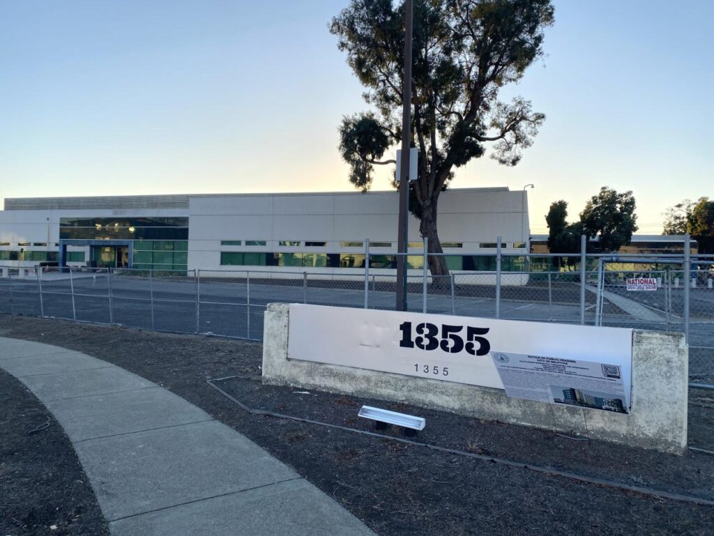 A one-floor industrial building and parking lot surrounded by a chain link fence at 1355 California Circle.