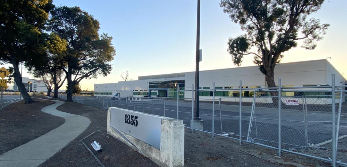 A one-floor industrial building and parking lot surrounded by a chain link fence at 1355 California Circle.