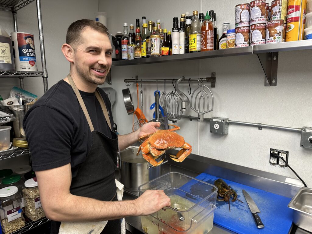 A man in a kitchen holds a crab over a basket.