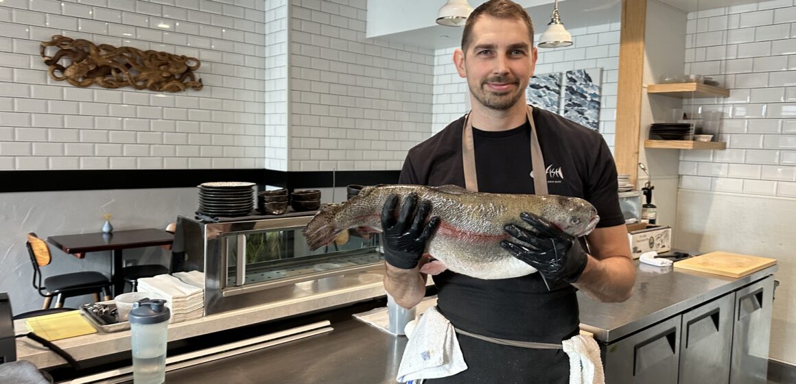 A man holding a fish stands behind the counter in a restaurant.