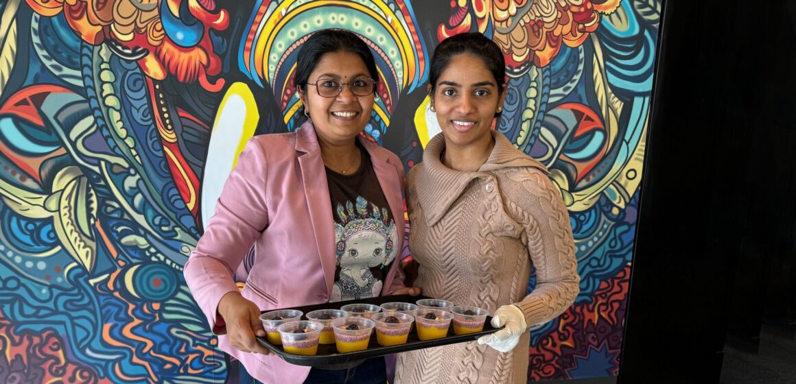 Two women stand facing the camera holding a tray of food.