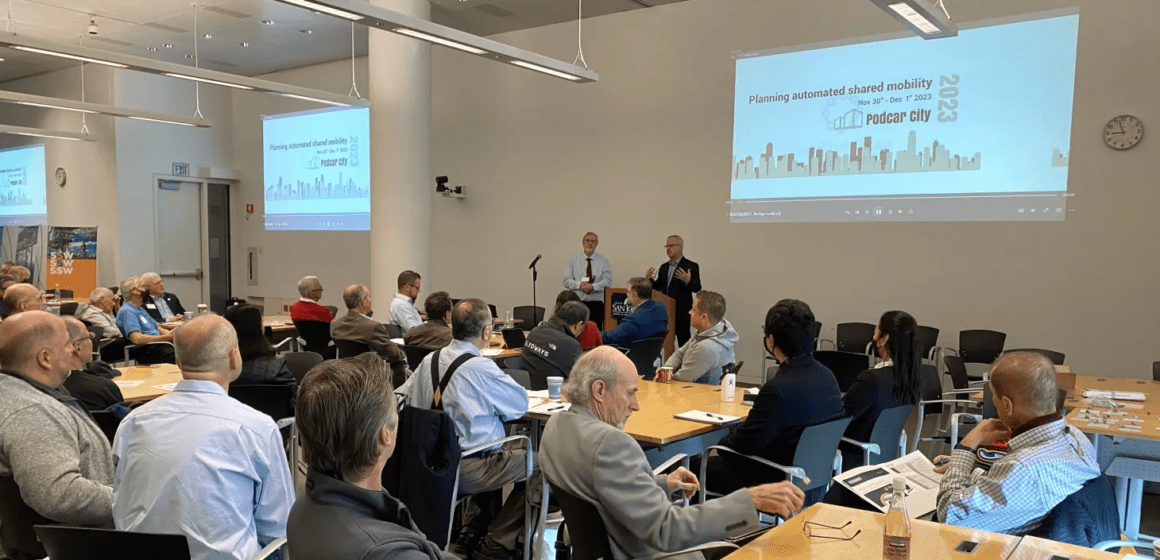 Crowd of people sit at tables in front of a speaker at the Podcar City 2023 conference.