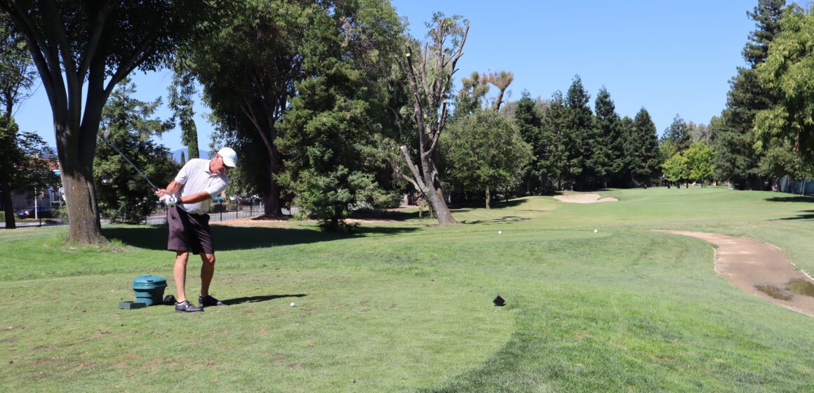 Tall man in white shirt and white hat swinging golf club on course