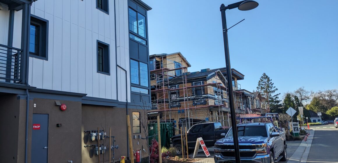 A finished white multi-story house next to a wooden house still under construction with scaffolding. In front of the houses is greenery and a pickup truck.