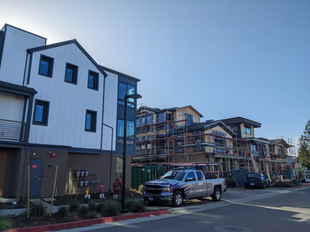 A finished white multi-story house next to a wooden house still under construction with scaffolding. In front of the houses is greenery and a pickup truck.