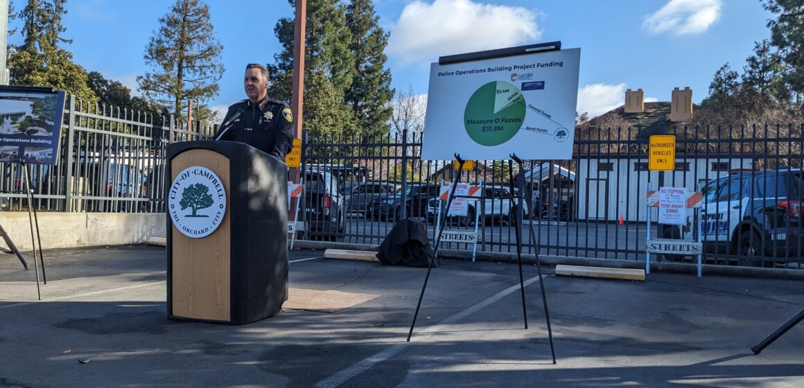 A police chief stands in front of a wooden podium with a microphone in a parking lot.