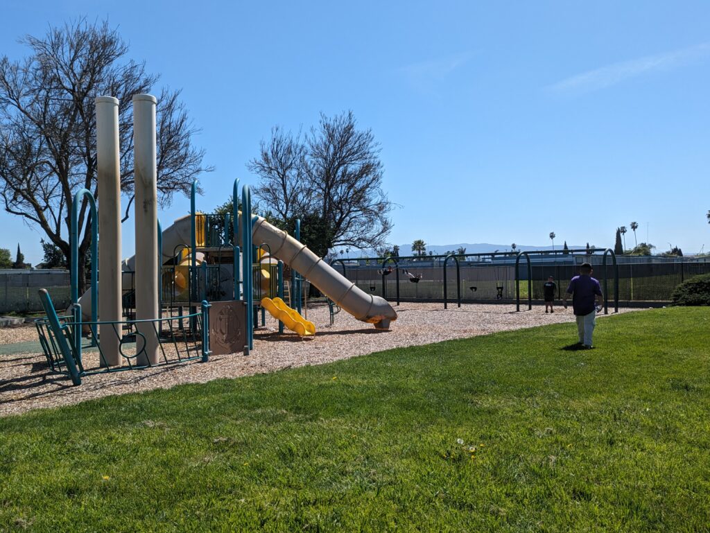 A photo of Sylvia Cassell Park in San Jose, where Councilmember Peter Ortiz said a pirate ship play structure used to be. Visible in photo: A play structure and a slide on tanbark with kids swinging in the background.