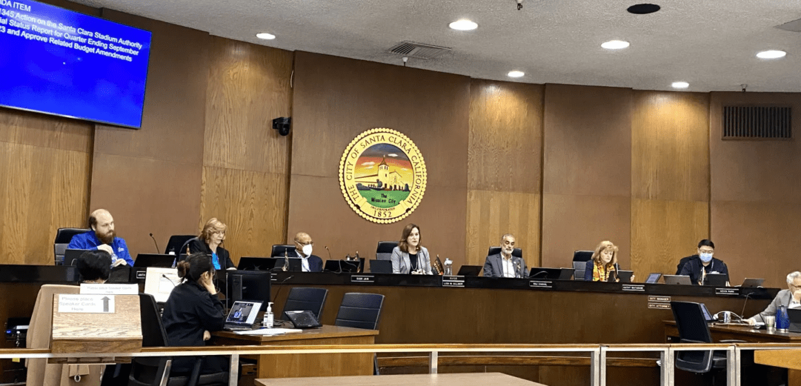 Seven people sitting on elevated dais with people working on desks in front of them