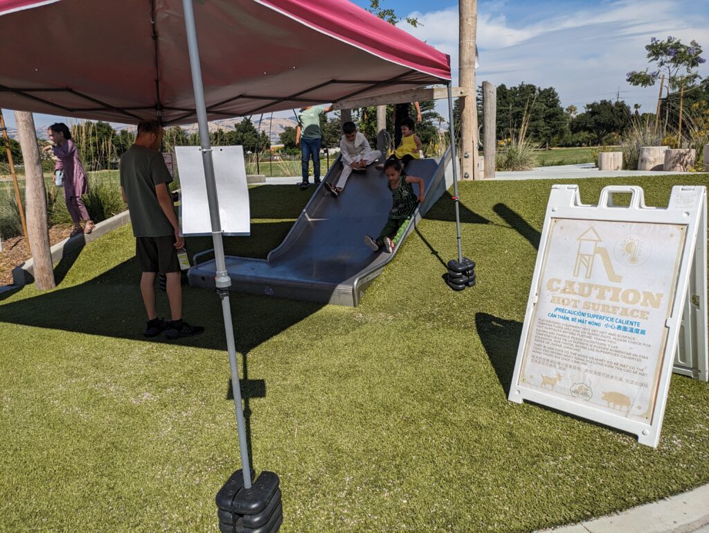 A pop-up canopy shades a small metal slide with children going down it. A sign warns families of the hot slide temperature.