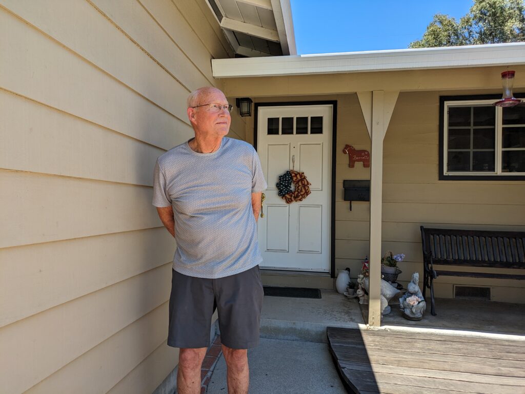 An older man wearing glasses stands in front of his front door