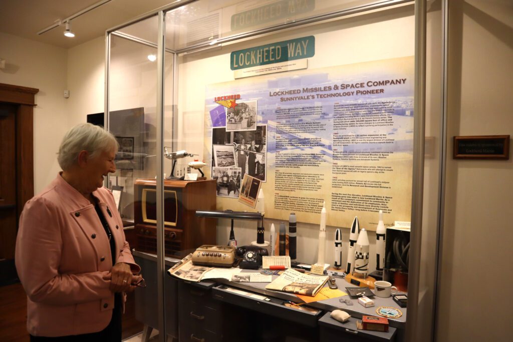 White haired woman in pink blazer examining the Lockheed display, which has model missiles and rockets, old phones, and newspaper clippings, all on top of a model engineer desk