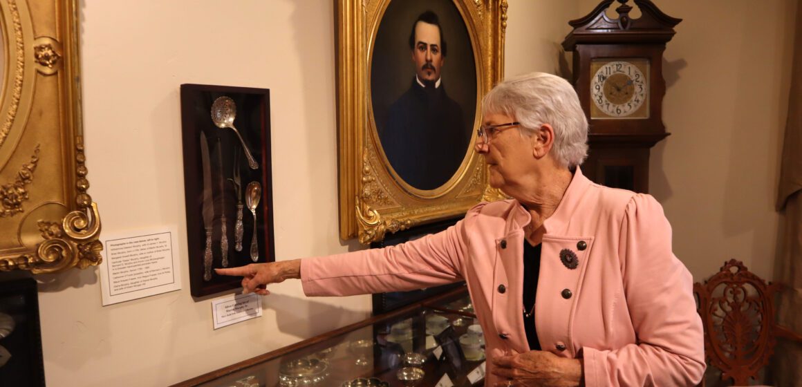 White haired woman in pink blazer pointing at silver cutlery mounted on museum wall, beside oil portrait and antique grandfather clock