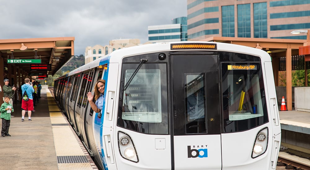 Woman waving out the driver's window of a new BART train while people wait on the platform