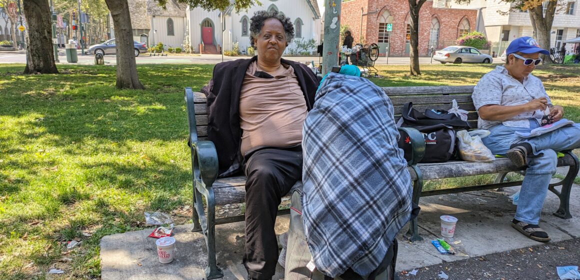 An unhoused man sits on a bench in a park