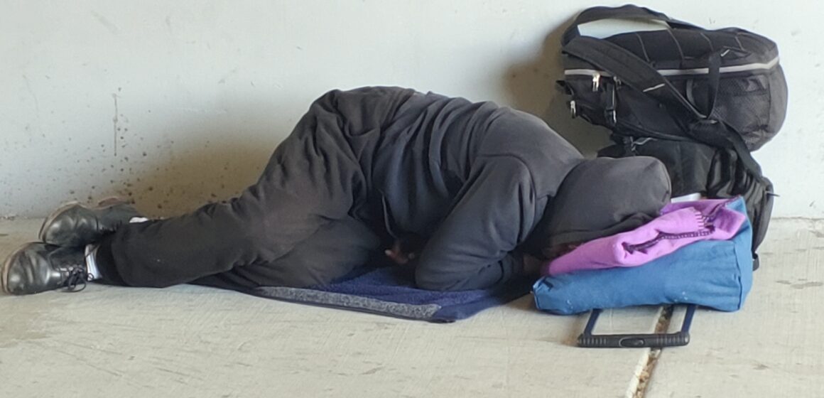 A homeless person sleeps on the ground under a highway overpass