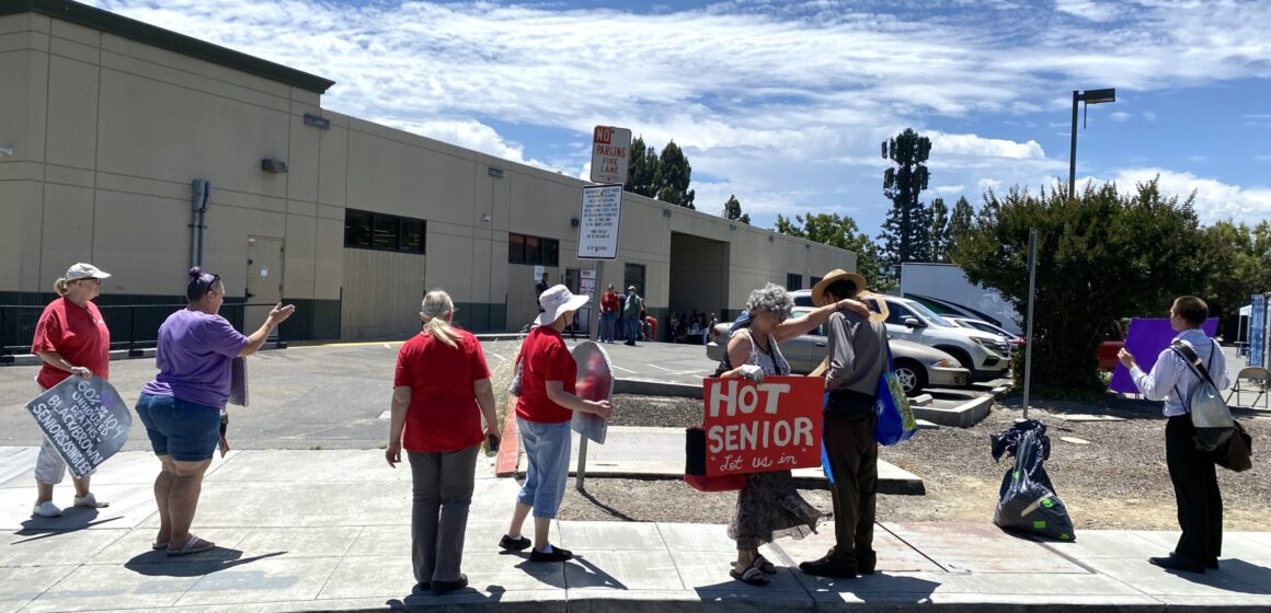 Group of people holding signs and standing on sidewalk near homeless shelter parking lot, with signs reading "60% of the 104 unhoused deaths — Black/Brown Seniors/Singles" and "Hot Senior, Let us in"