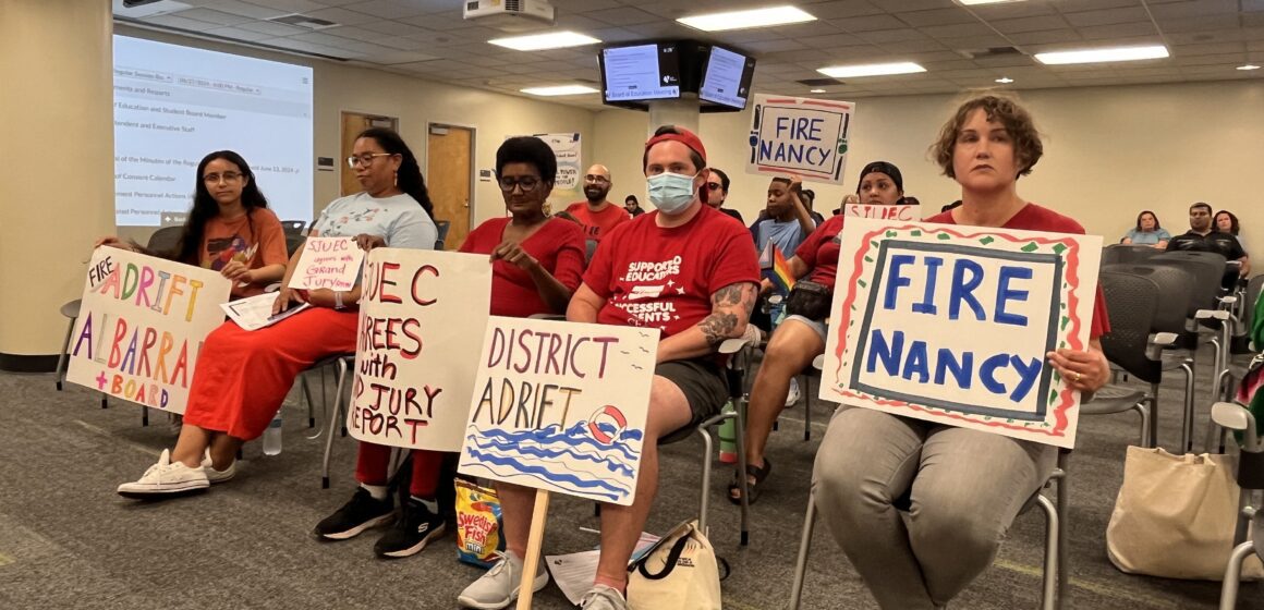 A row of people sitting in chairs holding protest signs at a school board meeting