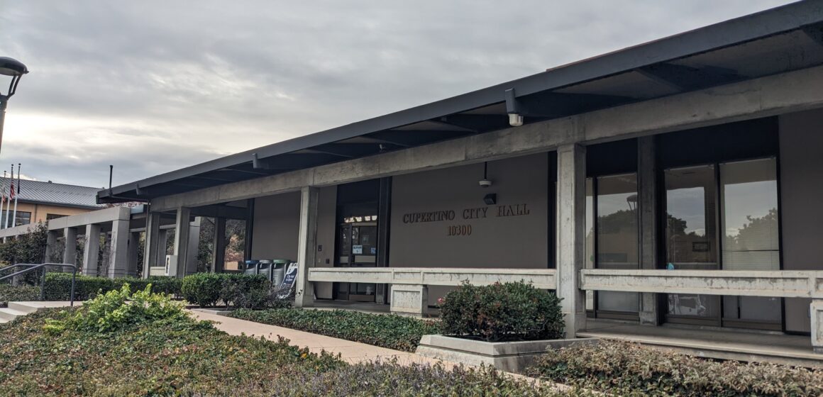 An exterior of an office building with the title Cupertino City Hall written on the front, behind green bushes.