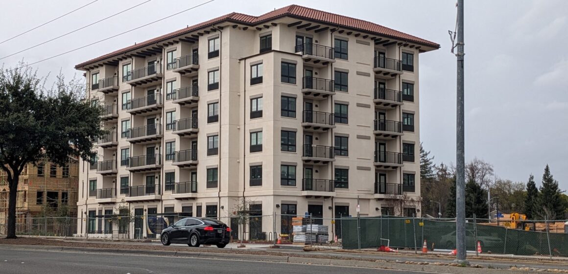 A six-story beige apartment building with a terracotta-colored roof sits behind an asphalt road.