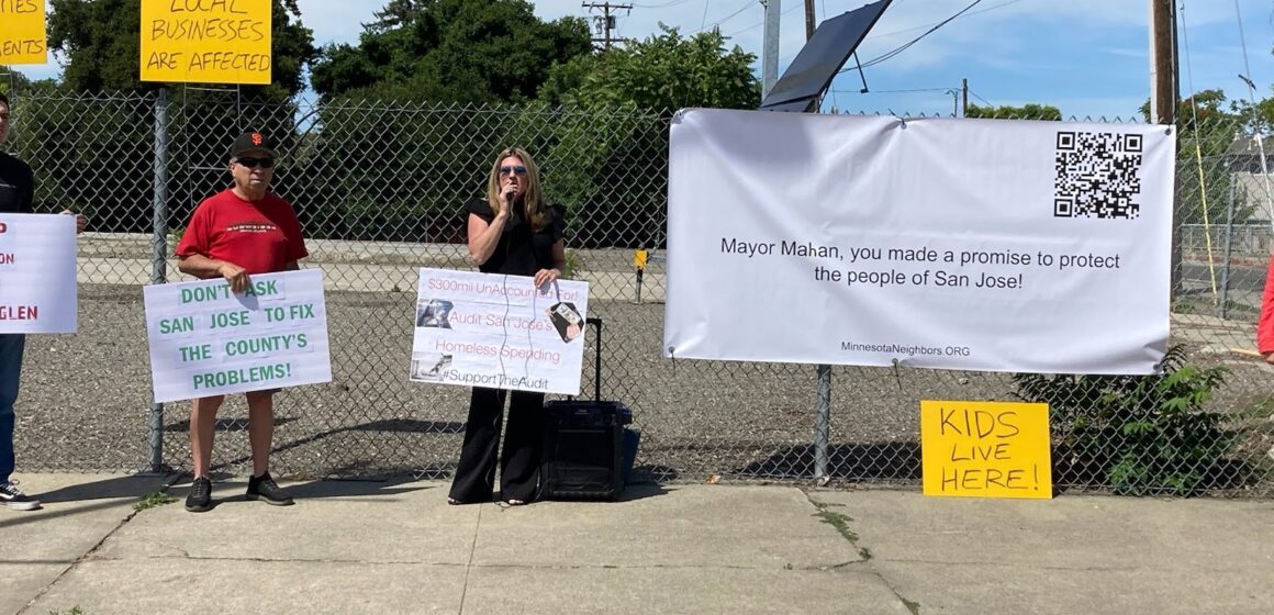 Three people standing in front of a chain link fence where a sign is hung reading "Mayor Mahan, you made a promise to protect the people of San Jose!" The person in the front, with long blonde hair and wearing black clothing, is holding a sign reading "$300mil Unaccounted For! Audit San Jose's Homeless Spending #SupportTheAudit" and another yellow sign beneath the hanging banner reads "Kids live here!"